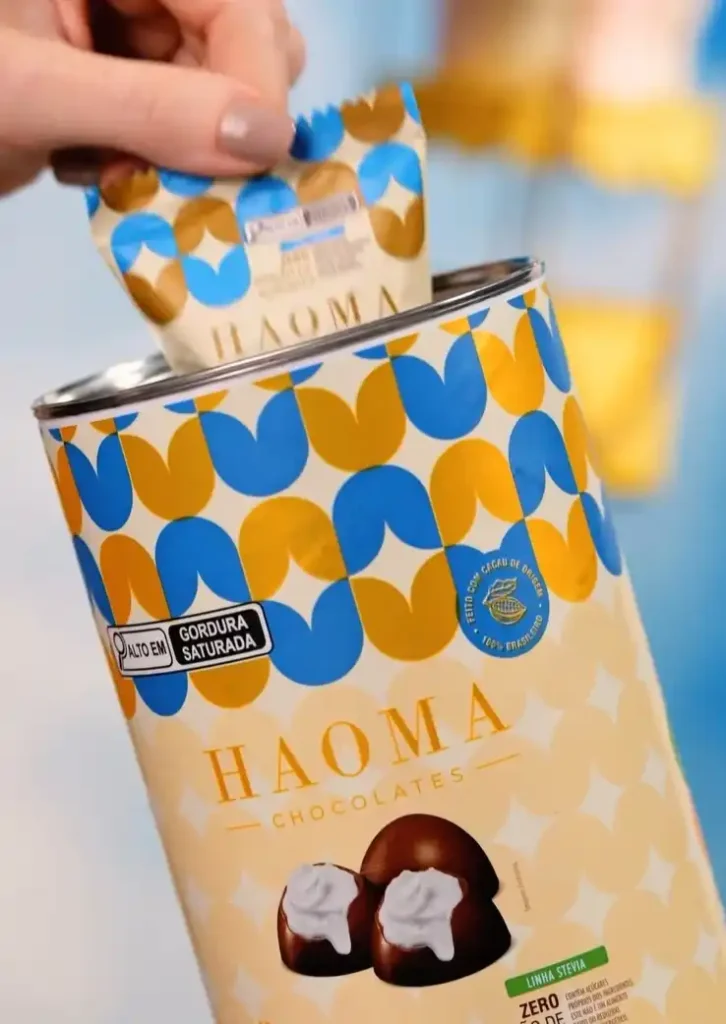 Uma lata alta de Haoma Chocolates com um padrão geométrico de círculos incompletos azuis e marrons. Uma mão está removendo um pequeno sachê individual da lata, que tem o mesmo design. A lata exibe o logo "HAOMA CHOCOLATES" em letras maiúsculas douradas, e abaixo dele, duas trufas de chocolate com cobertura branca. Há selos indicando "ALTO EM GORDURA SATURADA" e "FEITO COM CACAU DE VERDADE 100% BRASILEIRO". O fundo está desfocado em tons de azul claro e branco.