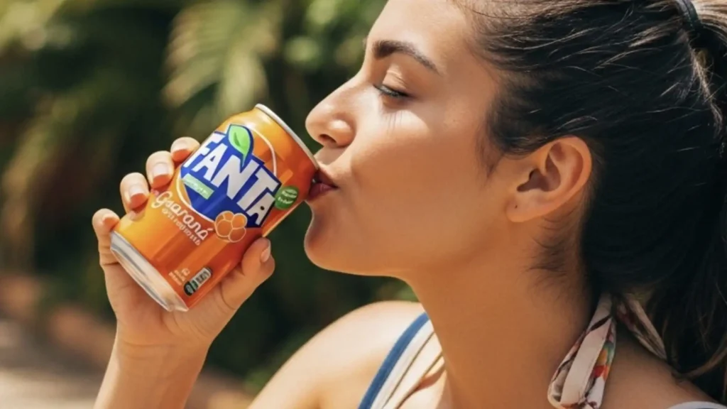 Uma mulher jovem e sorridente desfruta de uma Fanta Guaraná gelada em um dia ensolarado, com um fundo de folhagem tropical.