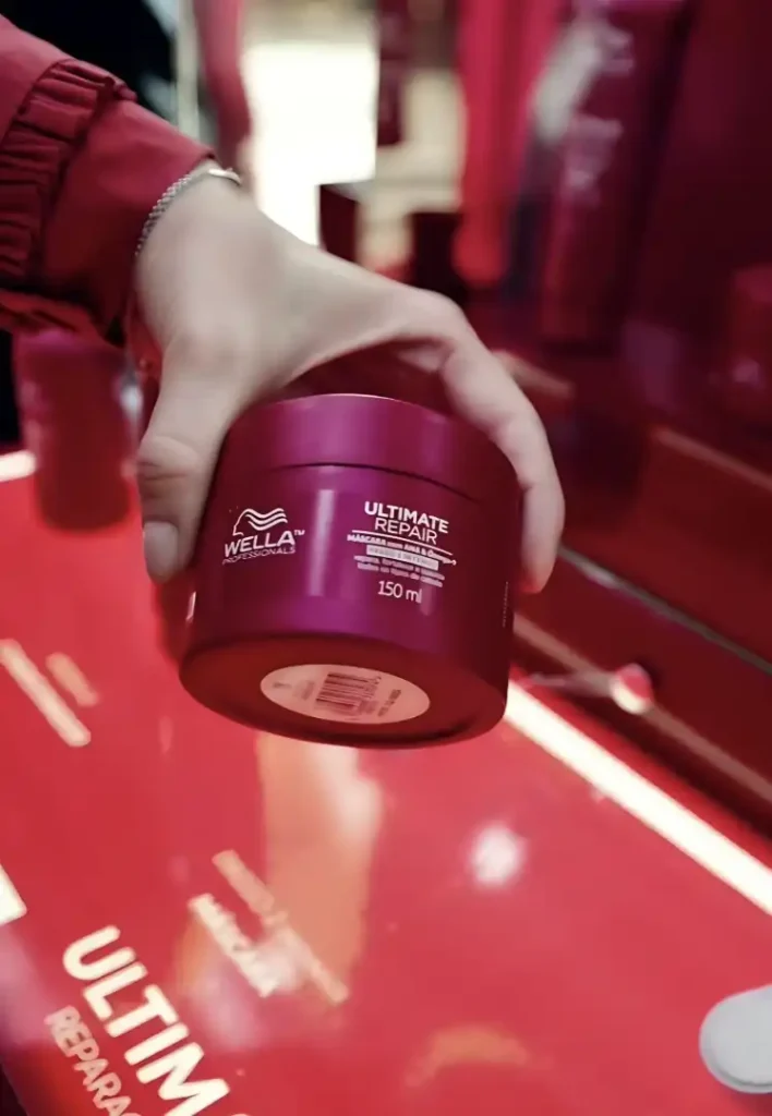 Uma pessoa segura um pote rosa de Wella Professionals Ultimate Repair Mask de 150 ml sobre uma superfície espelhada rosa/vermelha. O pote tem a tampa rosa escura e o corpo rosa claro com letras brancas. O rótulo diz "Wella Professionals" e "Ultimate Repair".