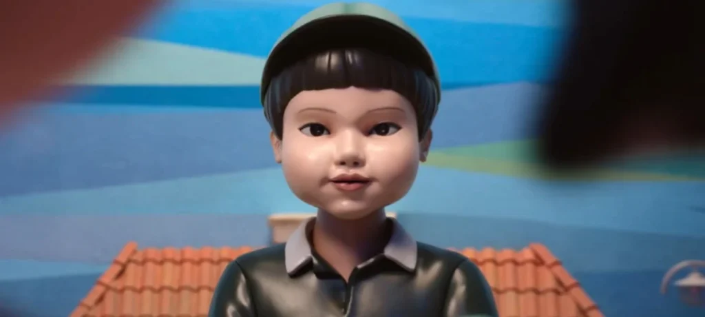 Uma close-up de um boneco sinistro de olhos arregalados, com pele pálida, cabelo escuro e liso, e uma expressão facial inexpressiva. O boneco usa um boné verde e uma camisa polo verde escura com colarinho branco. O fundo está desfocado, mas parece ser uma área externa com um telhado de terracota à vista.