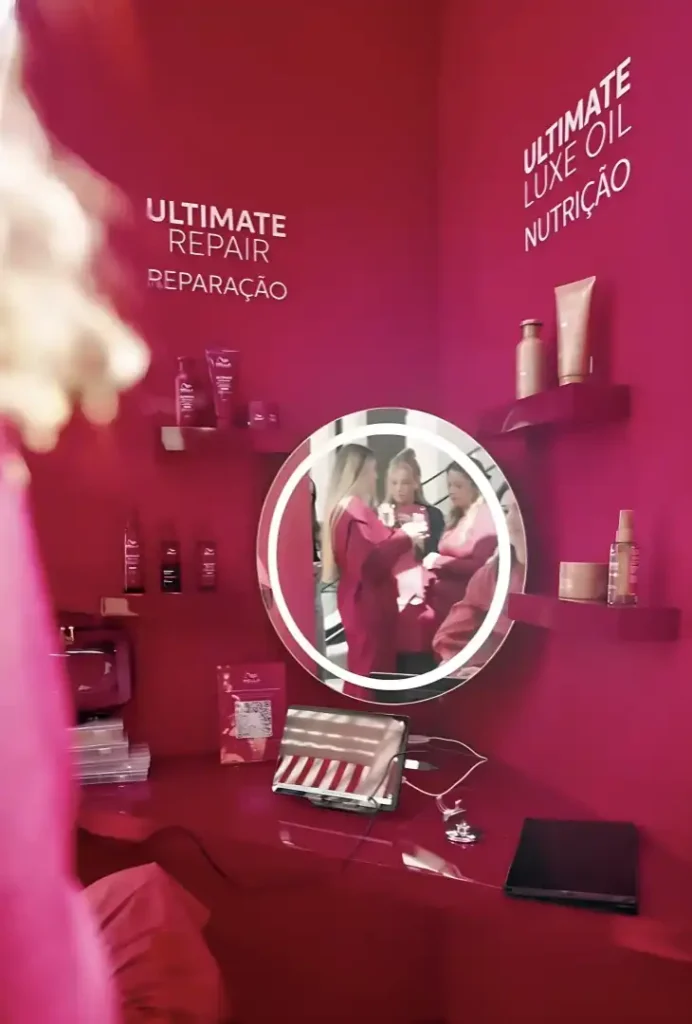 Uma cabine de beleza Wella Professionals Ultimate Luxe Oil com paredes cor-de-rosa intensas. O espaço é dedicado à linha "Ultimate Repair", focada em reparação, e à linha "Ultimate Luxe Oil", para nutrição. A cabine possui prateleiras com produtos da marca, um espelho redondo iluminado ao centro onde se refletem duas pessoas e uma bancada com um tablet e outros itens.