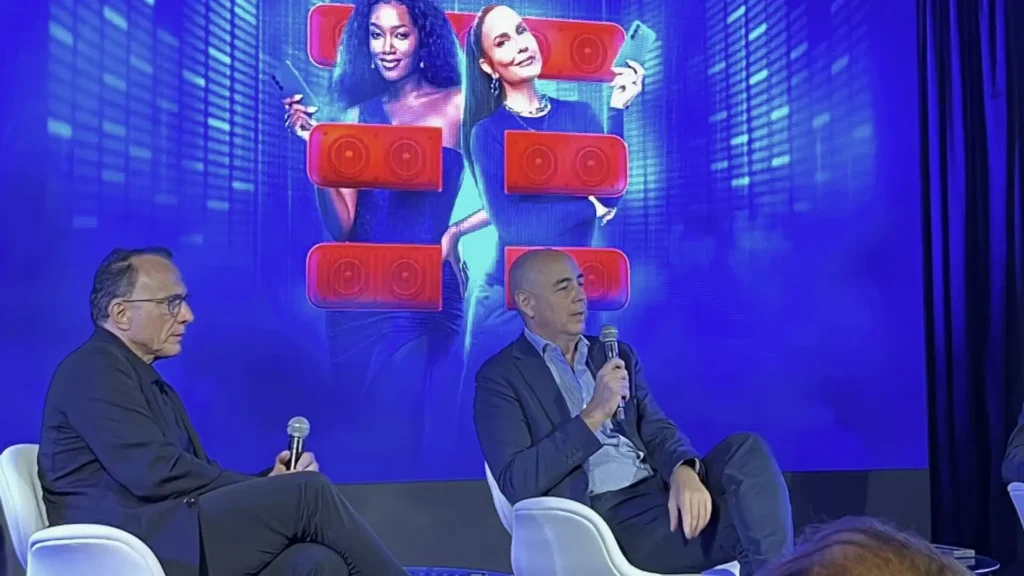 Duas mulheres sorridentes, uma com pele morena e outra com pele clara, seguram celulares com fundos azuis vibrantes. Ambas estão sentadas atrás de dois homens. O homem à esquerda está mais velho, usa óculos e veste uma camisa preta. Ele tem um microfone na mão. O homem à direita é mais jovem, com cabelo raspado e uma camisa azul escura. Ele também segura um microfone. Ambos os homens estão sentados em cadeiras brancas. No geral, o tema da imagem é azul com toques de vermelho.