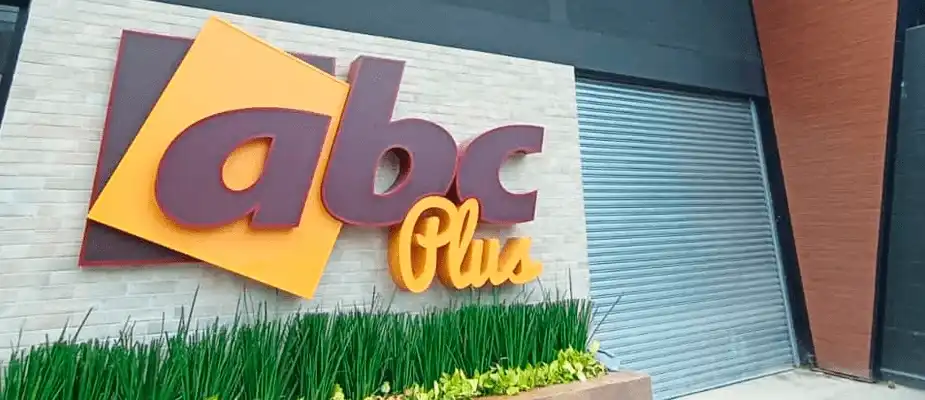 Uma foto em perspectiva de um sinal de loja montado na parede que diz "abc Plus". As letras "abc" são grandes e marrons, e a palavra "Plus" é menor e amarela, um pouco abaixo e à direita das letras "abc". As letras estão montadas em um fundo quadrado e retangular laranja. Abaixo do letreiro há uma plantadeira com grama verde. À direita do letreiro, há uma porta de enrolar cinza de metal que parece estar fechada.