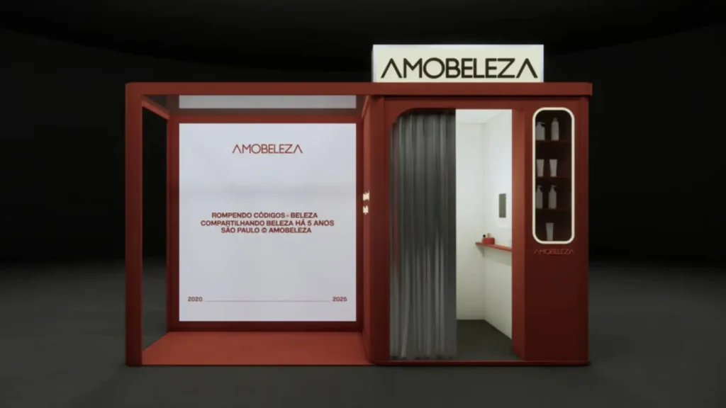 Uma representação digital 3D de um estande de beleza moderno. O estande tem uma estrutura vermelha escura e um interior branco. No lado esquerdo, há uma área aberta com uma grande tela branca exibindo o logotipo "AMOBELEZA" em letras pretas e as palavras "ROMPENDO CÓDIGOS - BELEZA COMPARTILHANDO BELEZA HÁ 5 ANOS SÃO PAULO @AMOBELEZA" e as datas "2020 2025" abaixo. O lado direito do estande é uma cabine fechada com uma cortina cinza escura na entrada e prateleiras vermelhas claras cheias de produtos de beleza brancos na parede interna. O logotipo "AMOBELEZA" também aparece na parte superior da estrutura e dentro da cabine. Um pequeno banner no canto superior esquerdo da imagem diz "Para sair do modo tela cheia, toque em Esc e pressione".