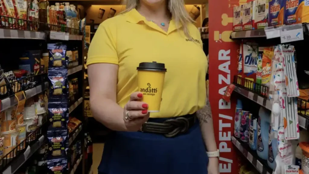 
Uma mulher loira vestindo uma camisa polo amarela e uma saia azul está em um corredor de supermercado, segurando um copo de café amarelo com a marca "andatti". Ela está de frente para a câmera e sorri ligeiramente. O corredor está cheio de prateleiras com produtos variados, incluindo lanches à esquerda e produtos de limpeza à direita. Um cartaz vermelho com os dizeres "PET/BAZAR" é visível no lado direito da imagem.