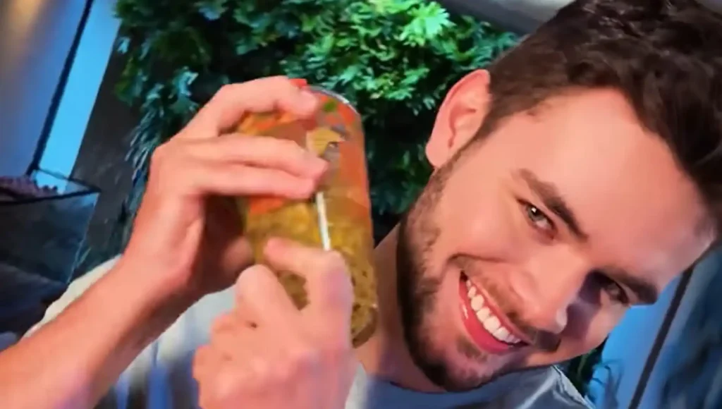 Um homem branco sorridente segura um pote de picles com a mão esquerda e uma faca com a direita, inserindo-a no pote. Ele tem cabelo castanho escuro e barba por fazer, e veste uma camiseta cinza. O fundo está desfocado com plantas e iluminação ambiente.