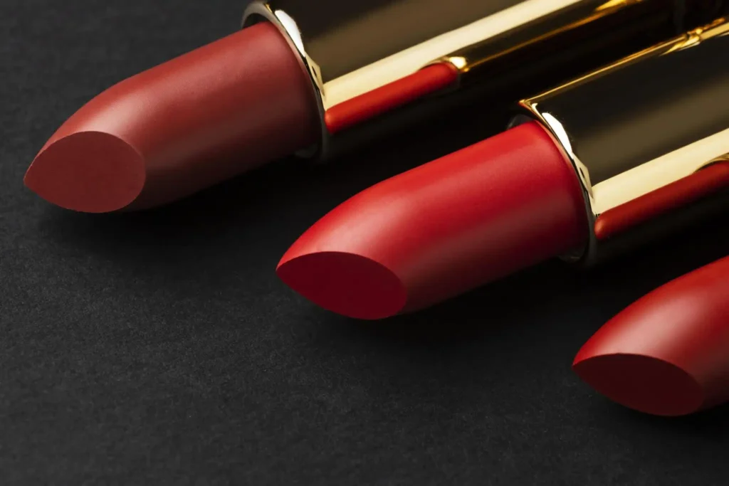 
Três batons em tons de vermelho e marrom, com embalagens douradas, dispostos lado a lado sobre uma superfície escura.