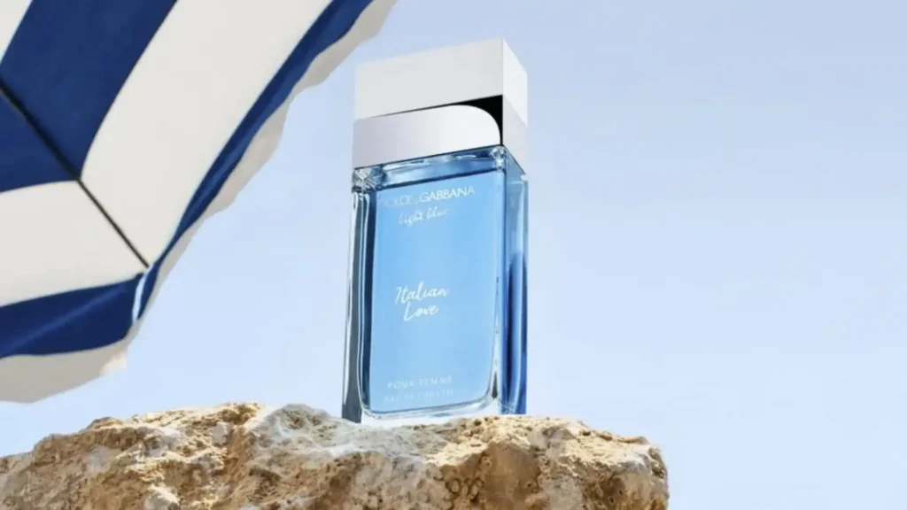 Uma garrafa retangular de perfume Dolce & Gabbana Light Blue Italian Love, na cor azul clara, está sobre uma rocha texturizada sob um guarda-chuva de praia listrado de azul e branco, com um céu azul claro ao fundo.