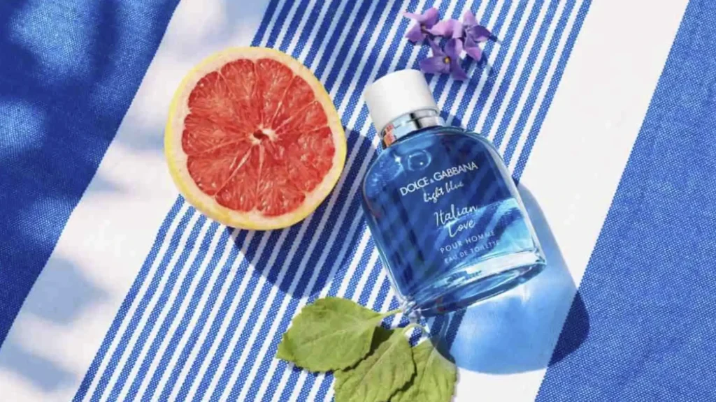 
Uma vista aérea de um frasco de perfume Dolce & Gabbana Light Blue Italian Love Pour Homme Eau de Toilette deitado sobre uma toalha de praia listrada de azul e branco. Ao lado do frasco há uma toranja cortada ao meio, um ramo de pequenas flores roxas e duas folhas verdes.