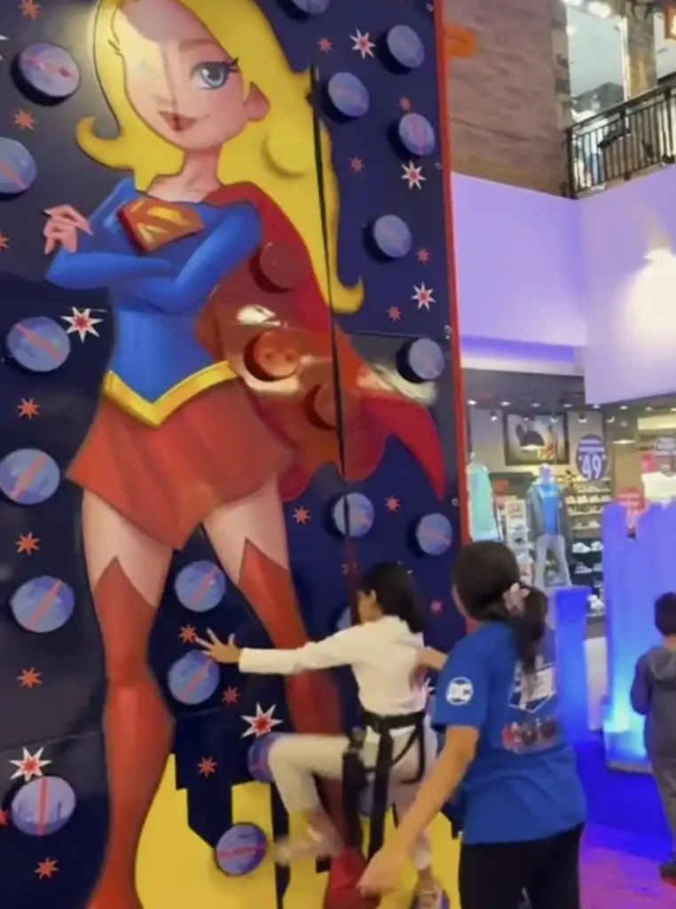 Uma menina escala uma parede de escalada colorida com a Supergirl em tamanho real pintada nela. A parede é predominantemente azul com padrões de estrela e círculos vermelhos e azuis servindo como apoios para as mãos. A Supergirl, com seu uniforme azul, vermelho e dourado, está retratada com os braços cruzados e uma capa esvoaçante. A menina está usando um arnês de segurança e uma camiseta branca de mangas compridas, e está concentrada em subir. Uma segunda pessoa, vestindo uma camiseta azul com um logotipo, está em pé na base da parede, aparentemente auxiliando a escaladora. No fundo, há outros elementos que sugerem um ambiente interno de recreação ou centro comercial.