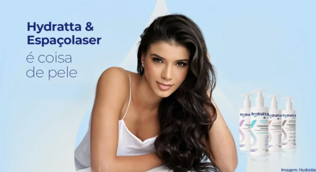 
Uma mulher com longos cabelos escuros, vestindo uma blusa branca de alças finas, olha para a frente com um leve sorriso. À esquerda dela, o texto "Hydratta & Espaçolaser é coisa de pele" está escrito em azul. À direita, três frascos brancos de produtos Hydratta estão alinhados. O fundo é um gradiente suave de azul claro.
