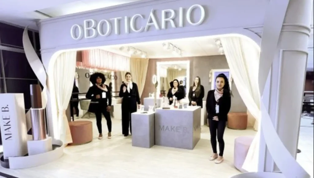 Uma imagem de um estande de vendas da marca "O Boticário", com a submarca "Make B." em destaque. O estande é elegante, com paredes claras e cortinas bege emoldurando a entrada. Dentro do estande, há prateleiras e displays com produtos de maquiagem. Cinco mulheres, aparentemente funcionárias do Boticário, estão posando em frente e dentro do estande. Elas estão vestindo roupas escuras (pretas ou tons escuros) e algumas usam colares claros. A mulher mais próxima à direita está sorrindo e olhando para a câmera. Há também um grande display em forma de batom com a marca "MAKE B." no lado esquerdo da imagem.