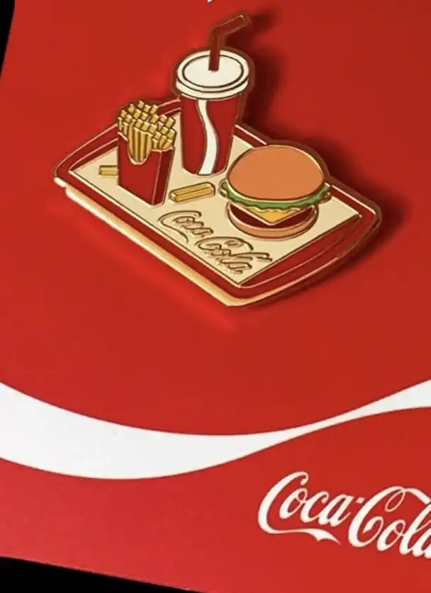 Um pin esmaltado em um fundo vermelho da Coca-Cola, mostrando uma bandeja dourada com a marca Coca-Cola escrita nela. Na bandeja há uma caixa vermelha de batatas fritas, um copo de Coca-Cola com um canudo e um hambúrguer com queijo e alface.