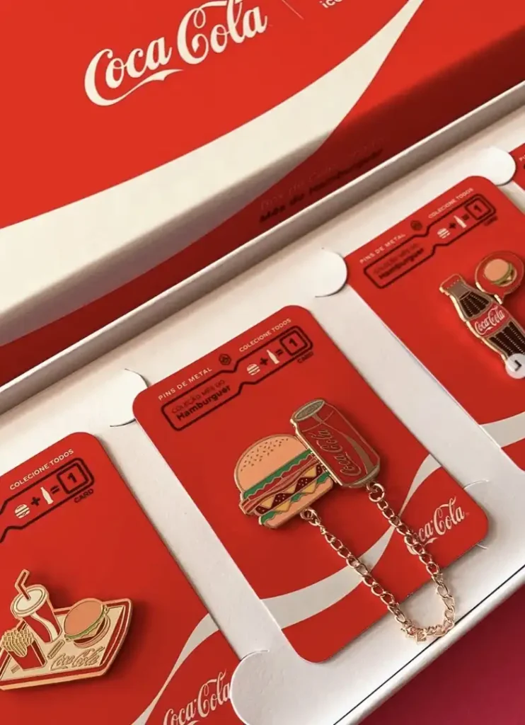 Uma coleção de pins esmaltados da Coca-Cola, apresentados em um estojo branco com tema da Coca-Cola. A imagem mostra três pins: um pin de hambúrguer com uma lata de Coca-Cola conectada por uma corrente, um pin de copo de Coca-Cola com batatas fritas e hambúrguer em uma bandeja, e um pin de garrafa de Coca-Cola. Todos os pins são montados em cartões vermelhos que detalham as instruções da coleção, insinuando que podem ser trocados por itens de menu.