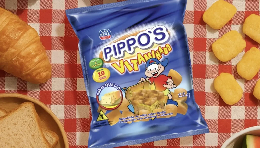 Uma sacola azul de "Pippo's Vitaminados" sabor queijo está no centro de uma toalha de mesa xadrez vermelha e branca. A sacola é adornada com desenhos de queijo e milho e uma mascote de desenho animado de um menino com um boné de beisebol, que parece estar segurando uma caixa de tesouro cheia dos salgadinhos. As palavras "ENRIQUECIDO COM 10 VITAMINAS E MINERAIS" e "22g" são visíveis na sacola. Ao redor da sacola, há salgadinhos dourados espalhados, um croissant e pão torrado em uma tigela de madeira.