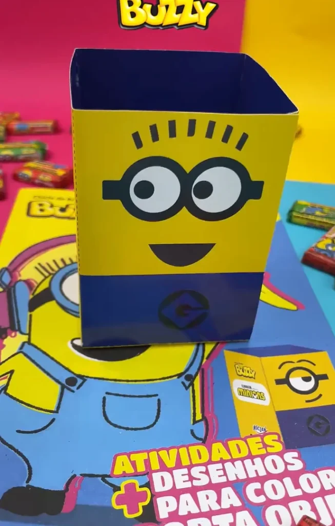 
Uma caixa amarela e azul em formato de porta-objetos com a ilustração de um Minion, com seus olhos grandes e redondos, boca sorridente e óculos pretos. A parte superior da caixa é azul escura e a inferior também é azul, com um símbolo circular dos Minions. A caixa está sobre um livro ou caderno também com tema dos Minions e em um fundo colorido com doces. A imagem parece ser de um produto infantil.