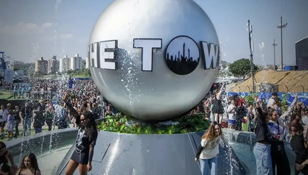 Uma fonte com um globo espelhado no centro tem a frase "THE TOWN" grafada em letras maiúsculas na metade do globo. A fonte está borrifando água e há pessoas reunidas ao redor.