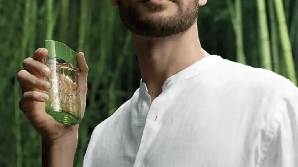 Um homem barbudo, usando uma camisa branca de linho, segura um frasco verde translúcido de desodorante colônia Arbo Puro. O fundo é composto por plantas verdes altas, que parecem ser bambu.