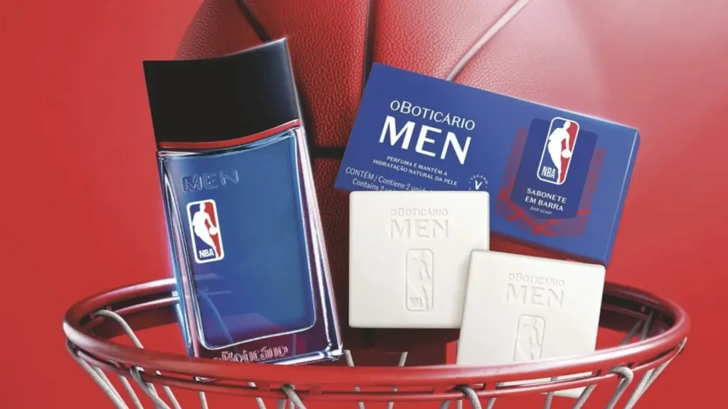 Uma garrafa azul e preta do perfume "O Boticário Men NBA" está inclinada contra uma bola de basquete laranja avermelhada em um aro de basquete vermelho e branco. Dois sabonetes brancos em barra "O Boticário Men NBA" estão posicionados à direita da garrafa, e a caixa azul escura para os sabonetes está atrás deles.