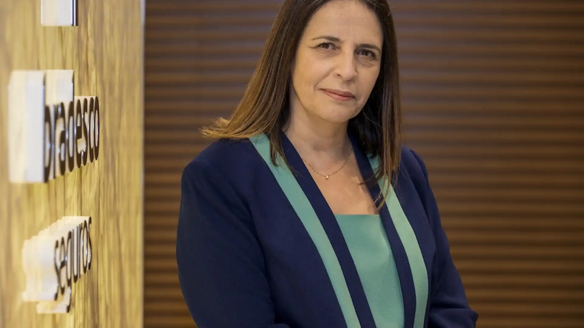Foto de Valdirene Soares Secato, diretora de Recursos Humanos, sorrindo gentilmente para a câmera. Ela usa um blazer azul-marinho sobre uma blusa azul-esverdeada. O fundo tem um painel de madeira com a marca 'Bradesco Seguros' em letras 3D brancas e douradas.