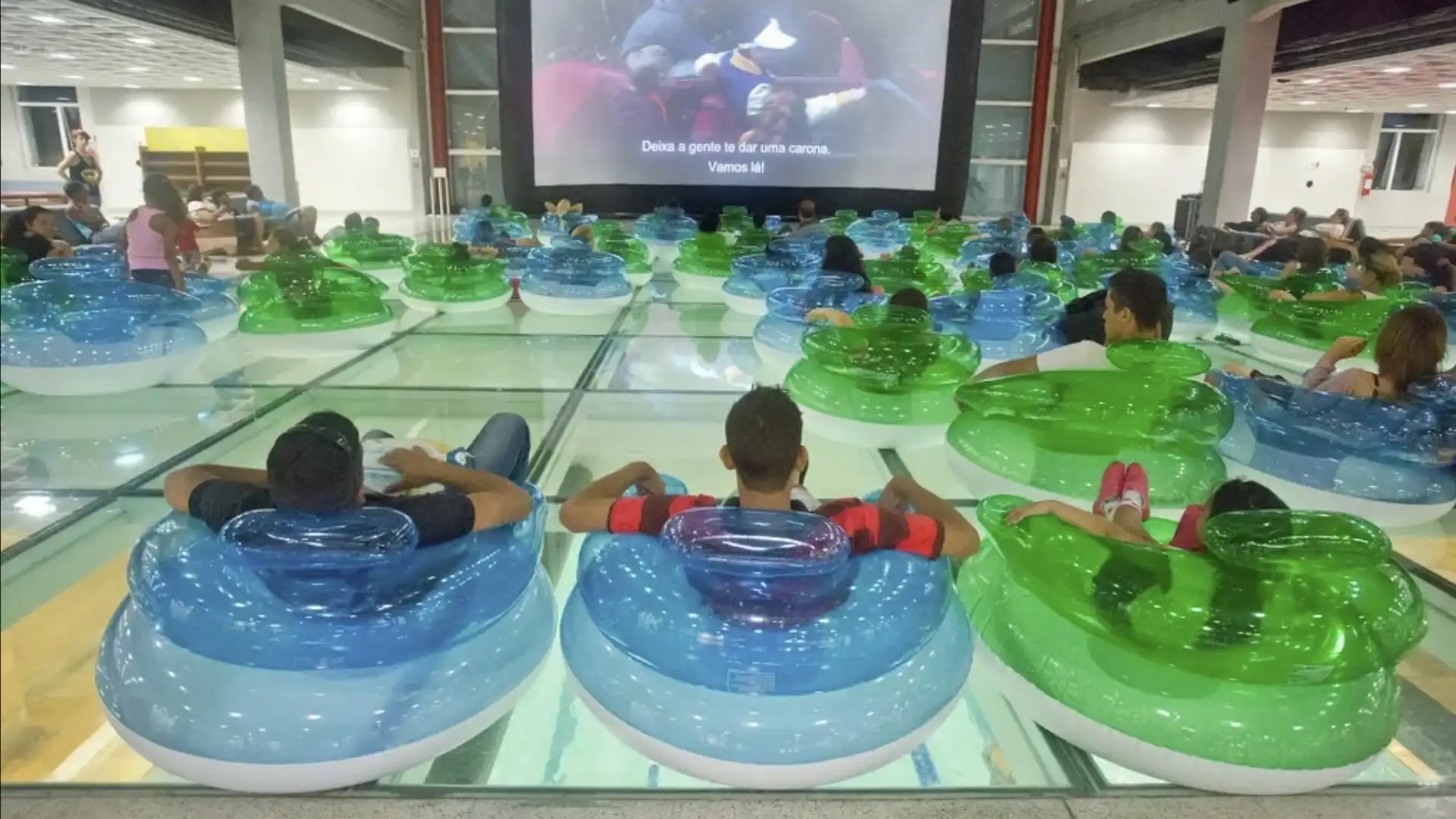 Pessoas sentadas em boias transparentes, dentro de uma piscina com fundo de vidro, assistindo a um filme.