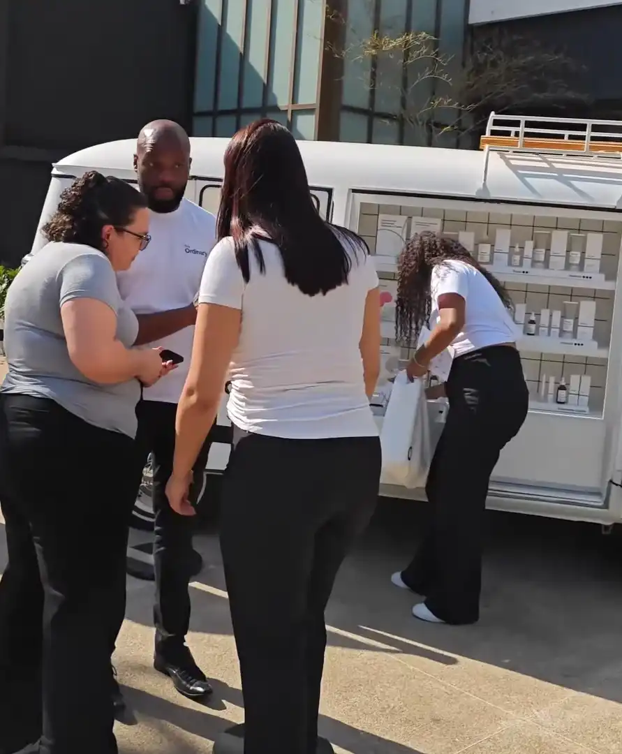 Profissionais de The Ordinary interagem com clientes em uma van de ativação da marca, expondo produtos para venda e demonstração.