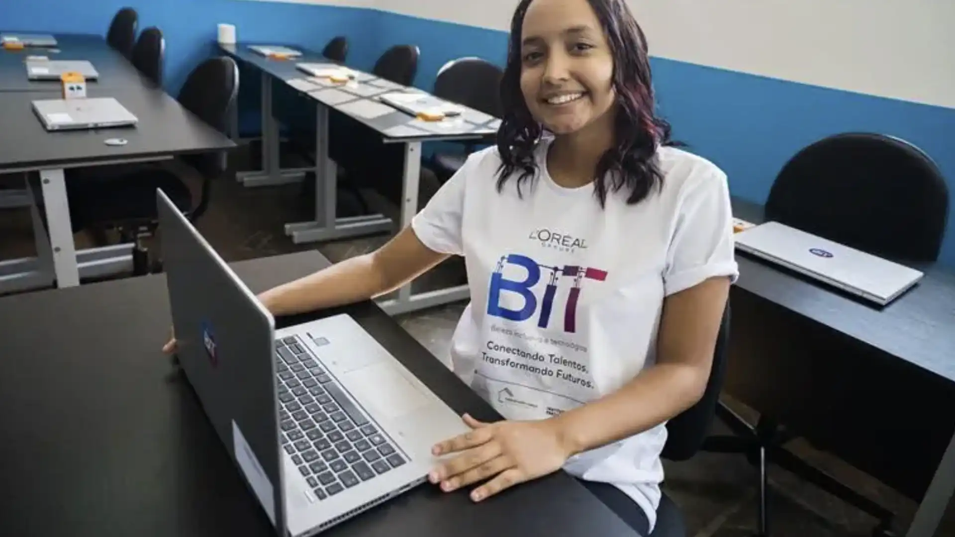 Um close de uma jovem brasileira sorrindo, sentada em uma mesa de aula com um notebook aberto.