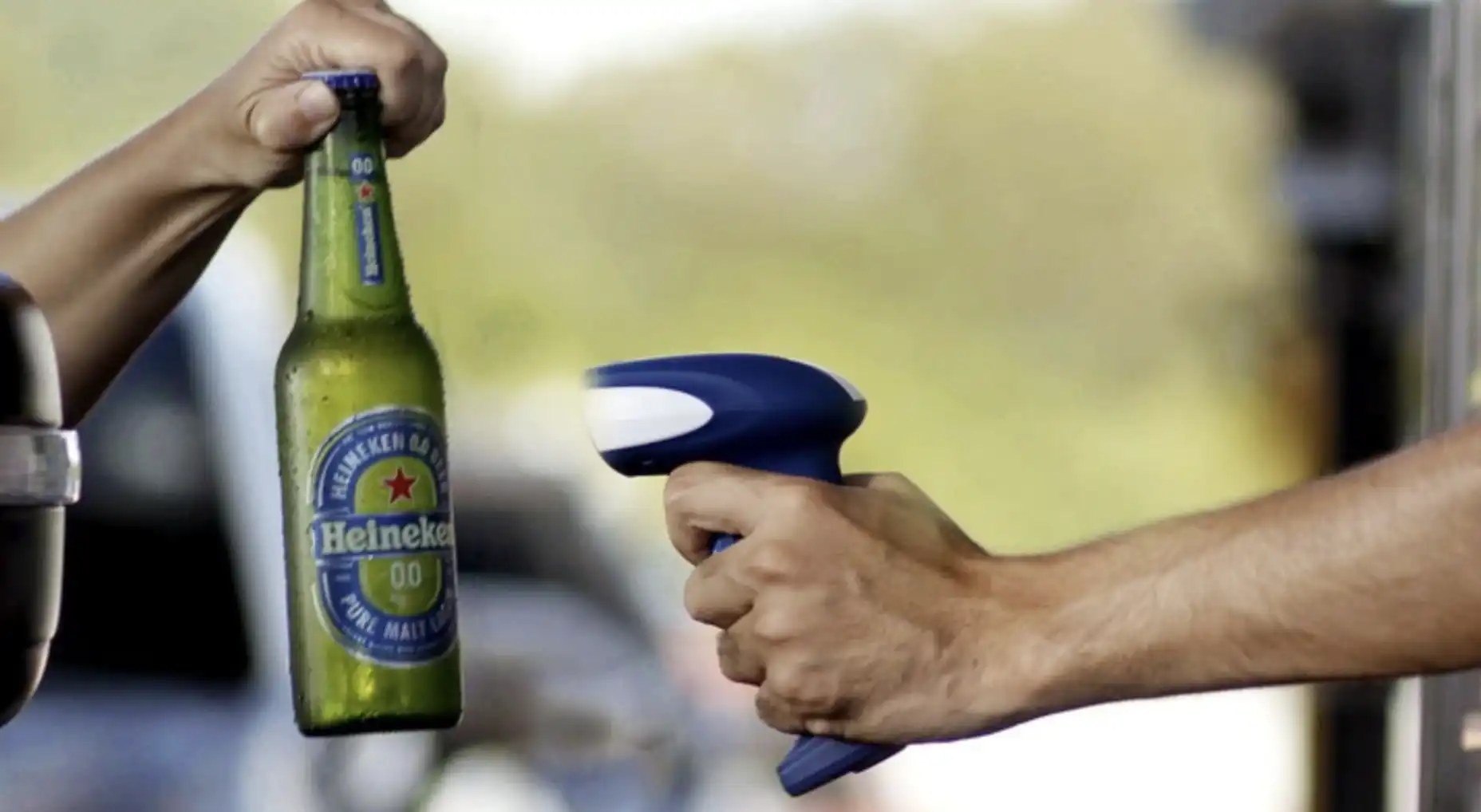 Duas mãos interagem, uma segura uma garrafa de Heineken 0.0, e a outra um scanner azul.