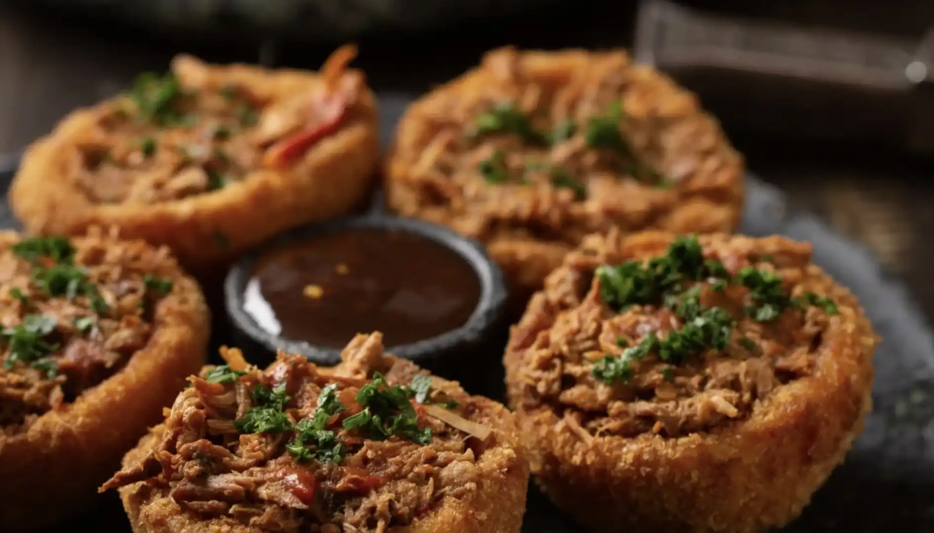 Seis porções de carne desfiada em cestas fritas, com um molho escuro no centro.