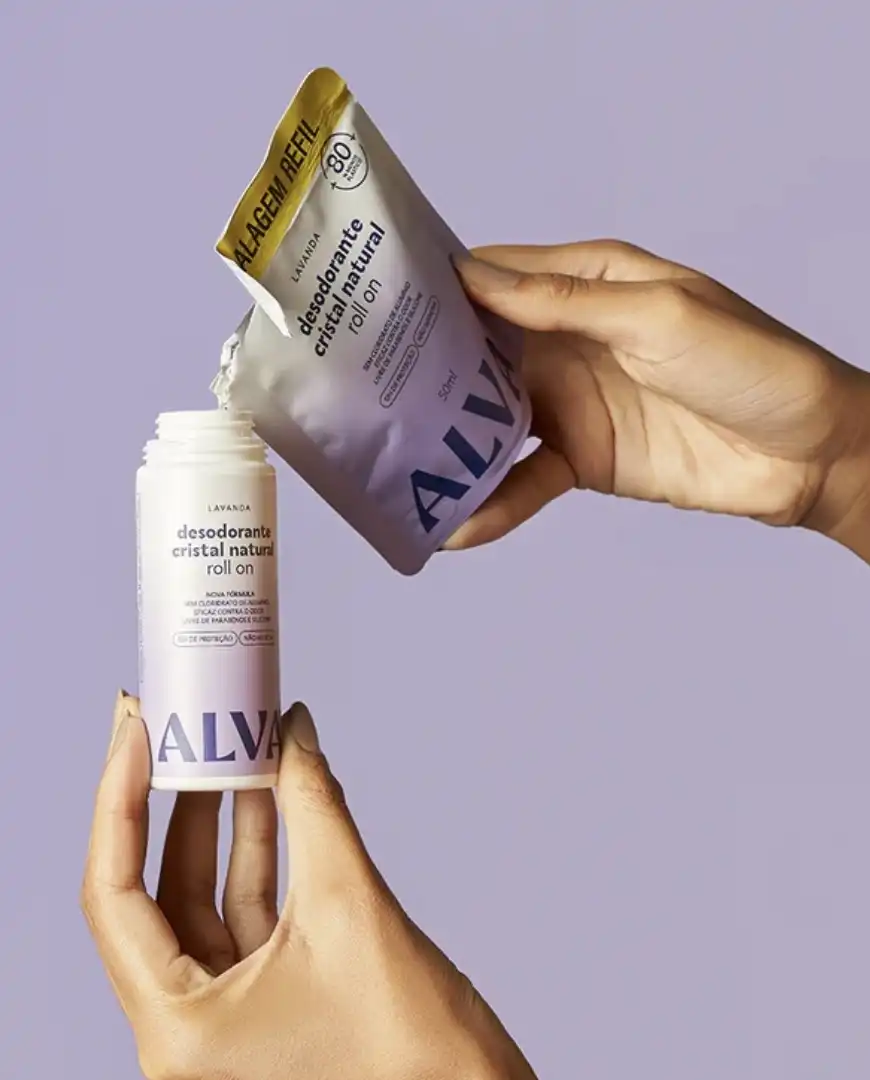 Mãos recarregando um frasco de desodorante roll-on natural de cristal da ALVA com um sachê de refil, em um fundo roxo suave.