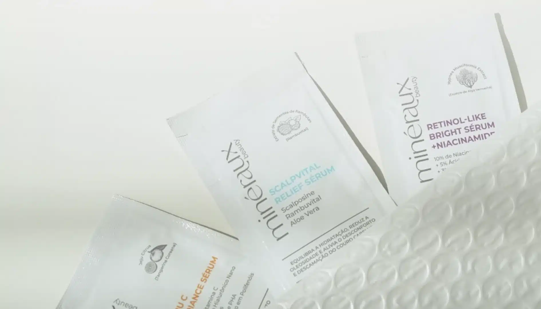Três sachês de amostras de produtos de beleza, da marca Mineralux Beauty, dispostos sobre uma superfície branca.