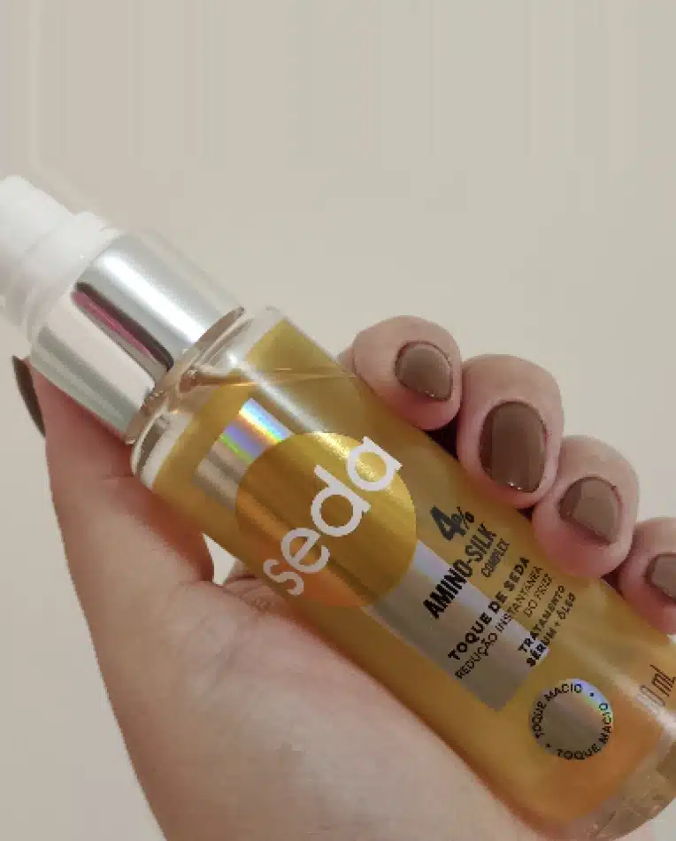 Pessoa segurando um frasco de SEDA Amino-Silk Complex com líquido amarelo. A embalagem é de vidro com tampa prateada e tem um rótulo com a marca e o nome do produto.