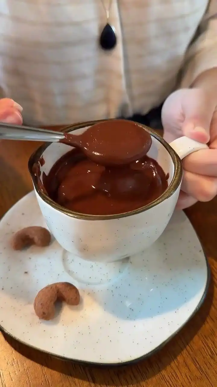 Uma pessoa segura uma xícara de porcelana branca com manchas, cheia de uma calda de chocolate espessa.