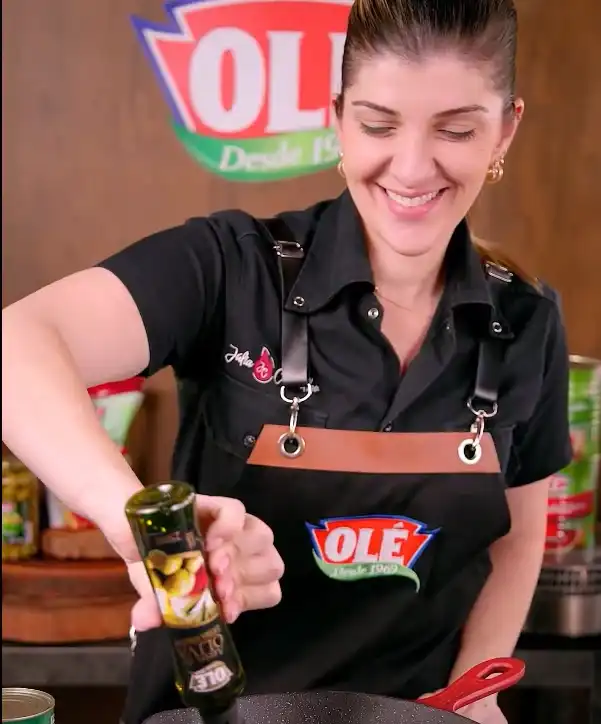 Uma mulher sorridente em um avental da marca Ole cozinha e adiciona azeite de uma garrafa.