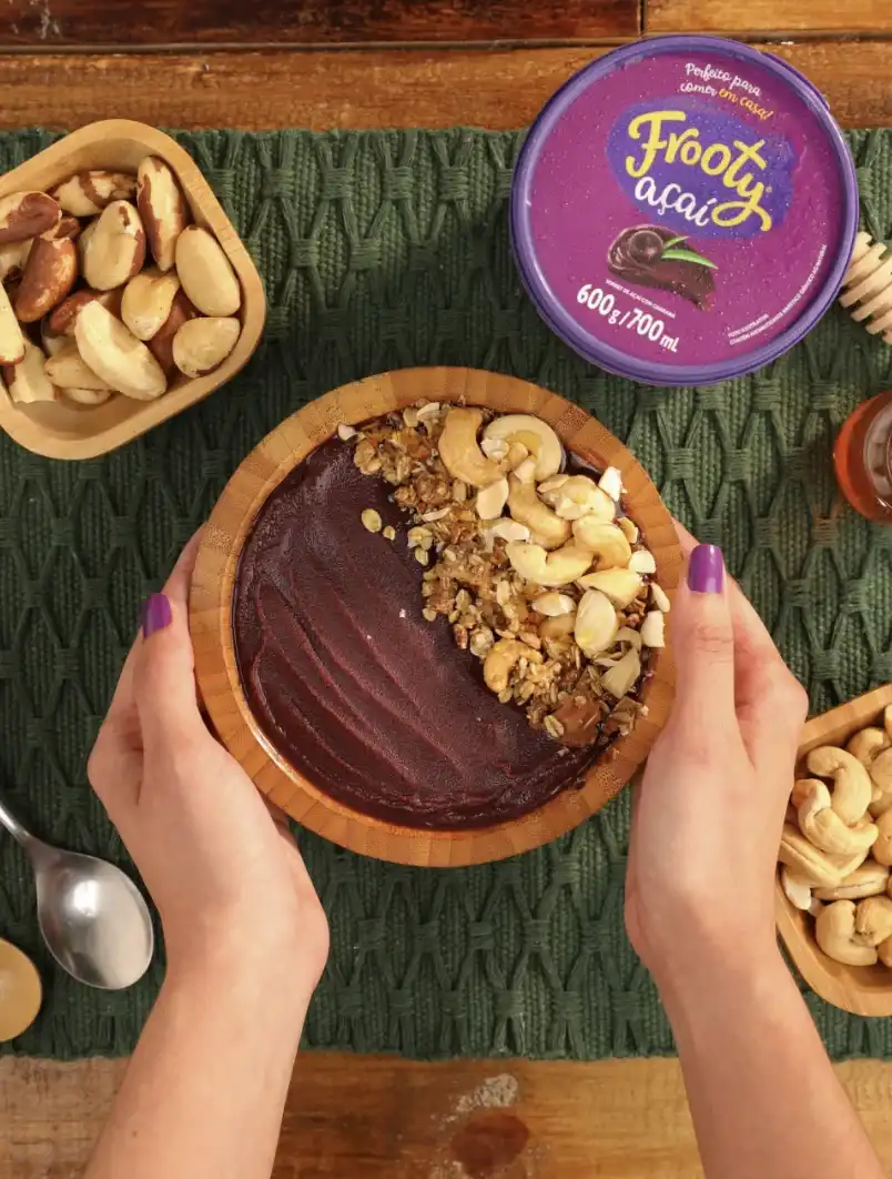 Uma pessoa segura uma tigela de açaí coberta com granola e castanhas de caju, vista de cima. A tigela é de madeira clara e está sobre uma toalha de mesa verde texturizada.