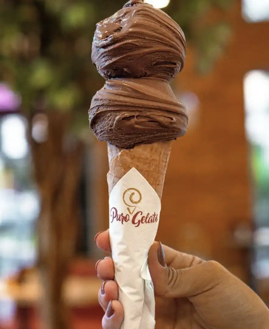 Uma pessoa segura uma casquinha com duas bolas de gelato de chocolate.
