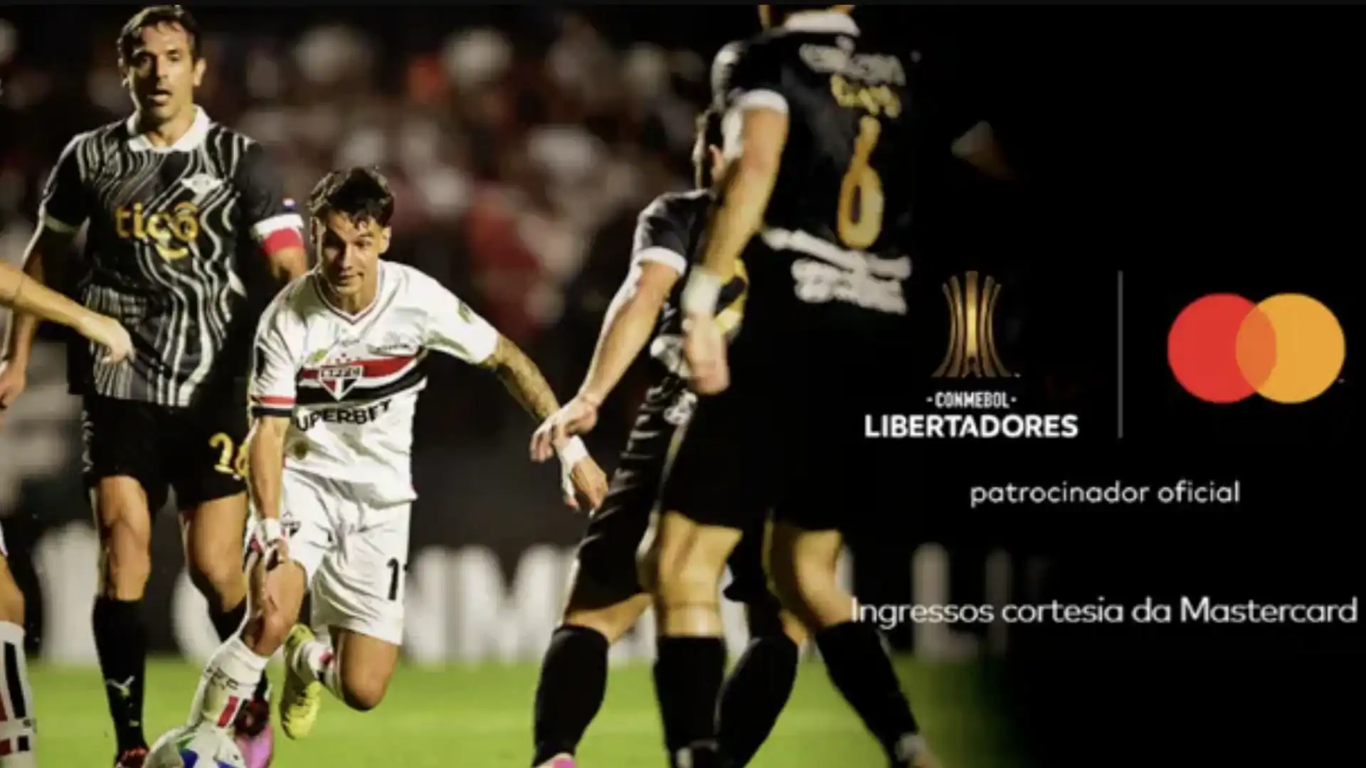 Uma bola de futebol em movimento, no canto de um campo em um estádio lotado de torcedores, sob a luz do sol.