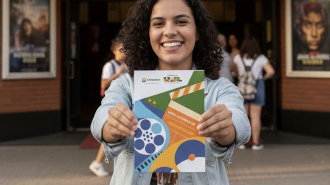 Mulher sorridente segura um álbum de figurinhas em frente à fachada iluminada do cinema 