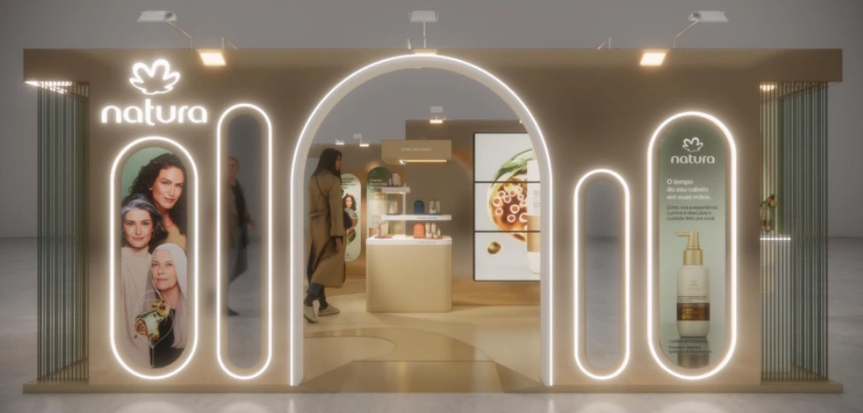 Fachada iluminada de um estande da Natura com arco central e painéis verticais exibindo modelos e produtos.