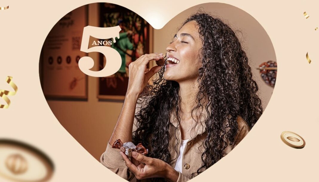 Mulher de cabelo cacheado comendo uma trufa de chocolate, segurando outra trufa embrulhada, dentro de um formato de coração com o número 
