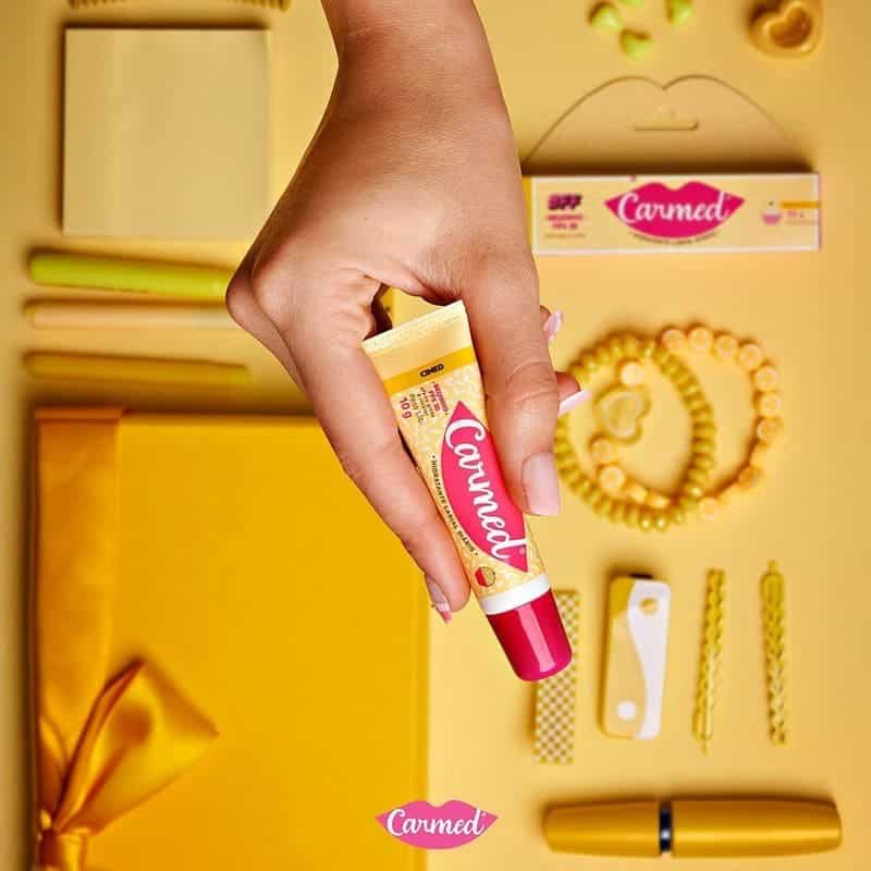 Uma mão com unhas rosadas segura um tubo amarelo e vermelho do hidratante labial Carmed sabor Cereja sobre um fundo amarelo vibrante, cercado por acessórios e itens de papelaria amarelos e em tons pastel.