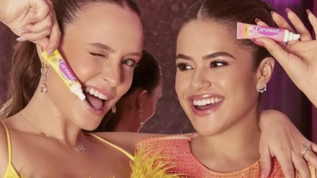 Duas jovens celebridades brasileiras, Larissa Manoela e Maisa, sorriem enquanto seguram tubos do hidratante labial Carmed em uma campanha publicitária colorida.