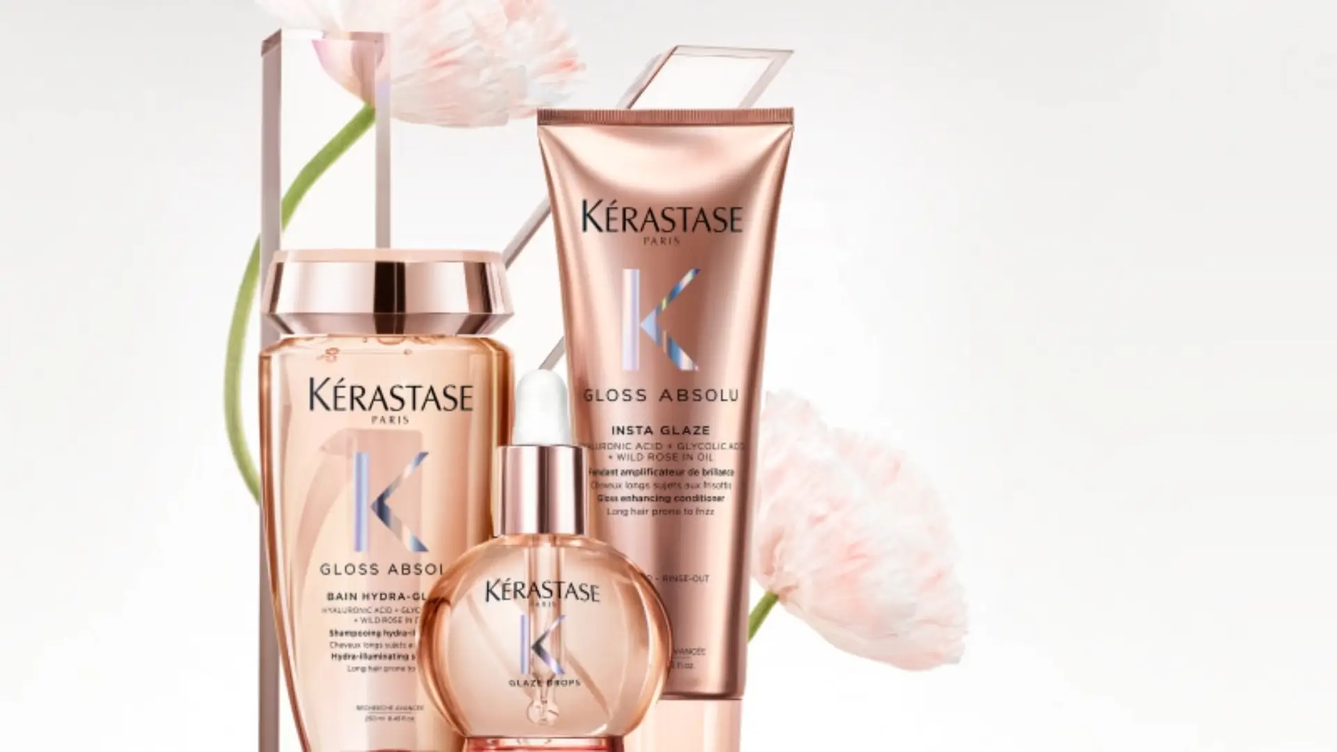 Close-up da linha de produtos capilares Kérastase Gloss Absolu, mostrando três embalagens em tom de cobre-rosado: um shampoo (Bain Hydra-Gloss), um óleo ou sérum (Glaze Drops) e um condicionador ou máscara (Insta Glaze). O fundo tem um desfoque suave com elementos florais rosados.