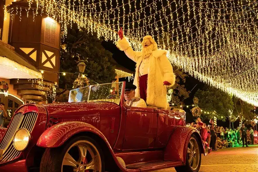 Papai Noel em um carro conversível vermelho antigo, acenando sob uma cobertura de luzes de Natal.