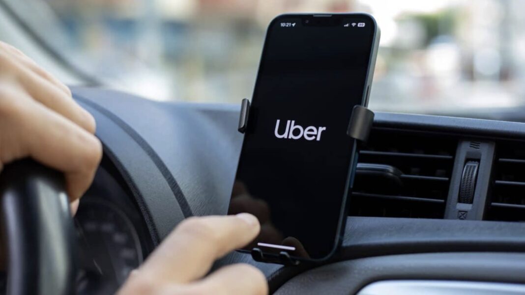 Mão de motorista toca na tela de um celular com o aplicativo Uber aberto, fixado em um suporte no painel do carro.Mão de motorista toca na tela de um celular com o aplicativo Uber aberto, fixado em um suporte no painel do carro.