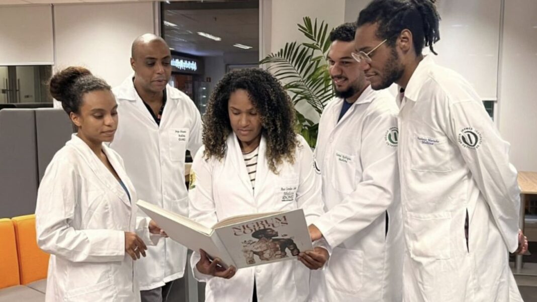 Cinco profissionais da saúde afro-brasileiros em jalecos brancos, reunidos, folheando juntos o livro 
