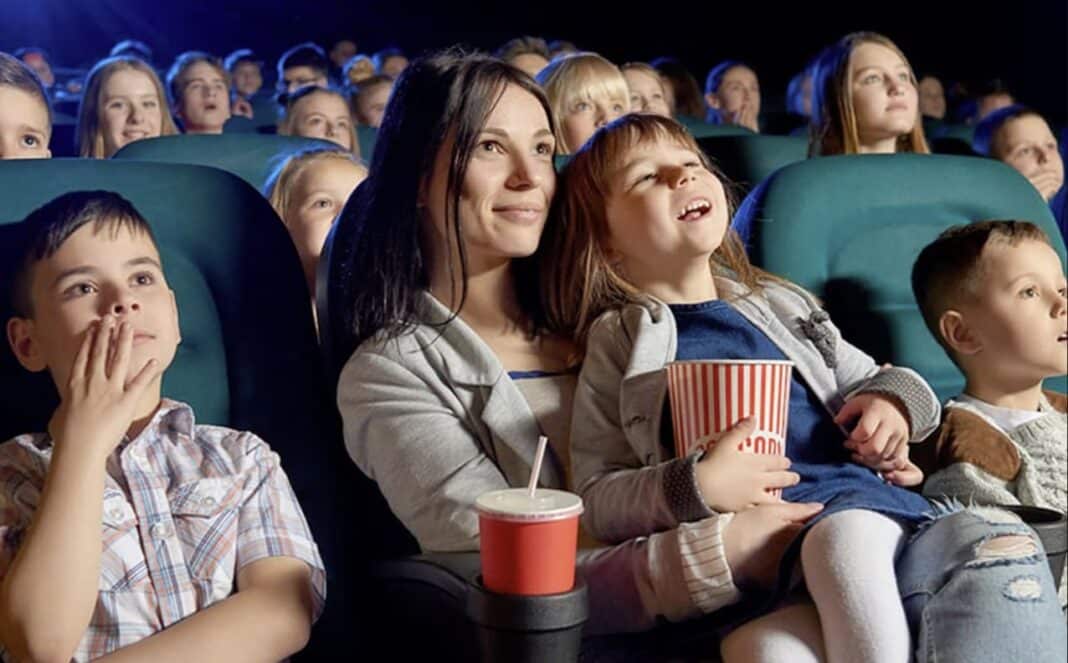 Mãe e filha assistindo a um filme no cinema com um balde de pipoca, cercadas por outras pessoas.
