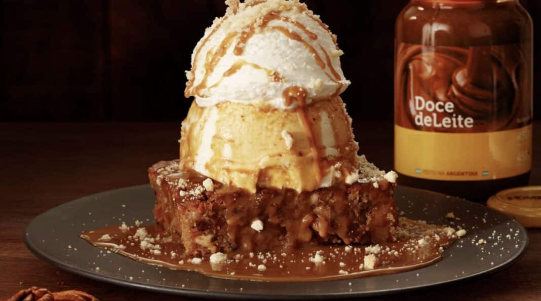 Sobremesa Havanna Thunder do Outback com brownie úmido coberto por doce de leite, sorvete de baunilha e chantilly, servida em um prato escuro com pote de doce de leite ao fundo.