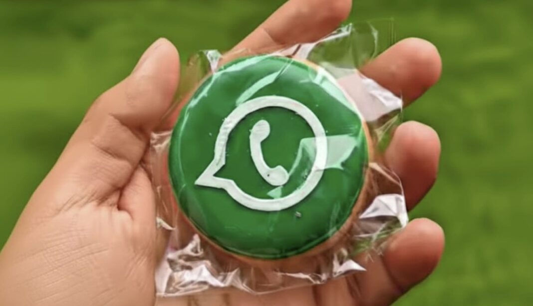 Biscoito redondo verde do WhatsApp embalado sendo segurado por uma mão, com fundo de grama sintética verde.