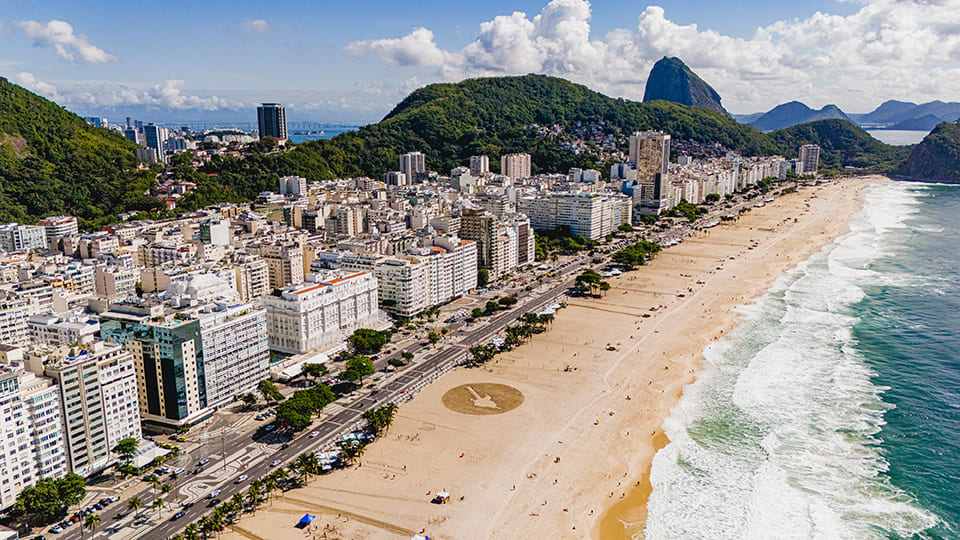 Vista aérea panorâmica da Praia de Copacabana, Rio de Janeiro, com a cidade e o Pão de Açúcar ao fundo, e uma arte na areia em formato de guitarra promovendo o Rock in Rio 2026.